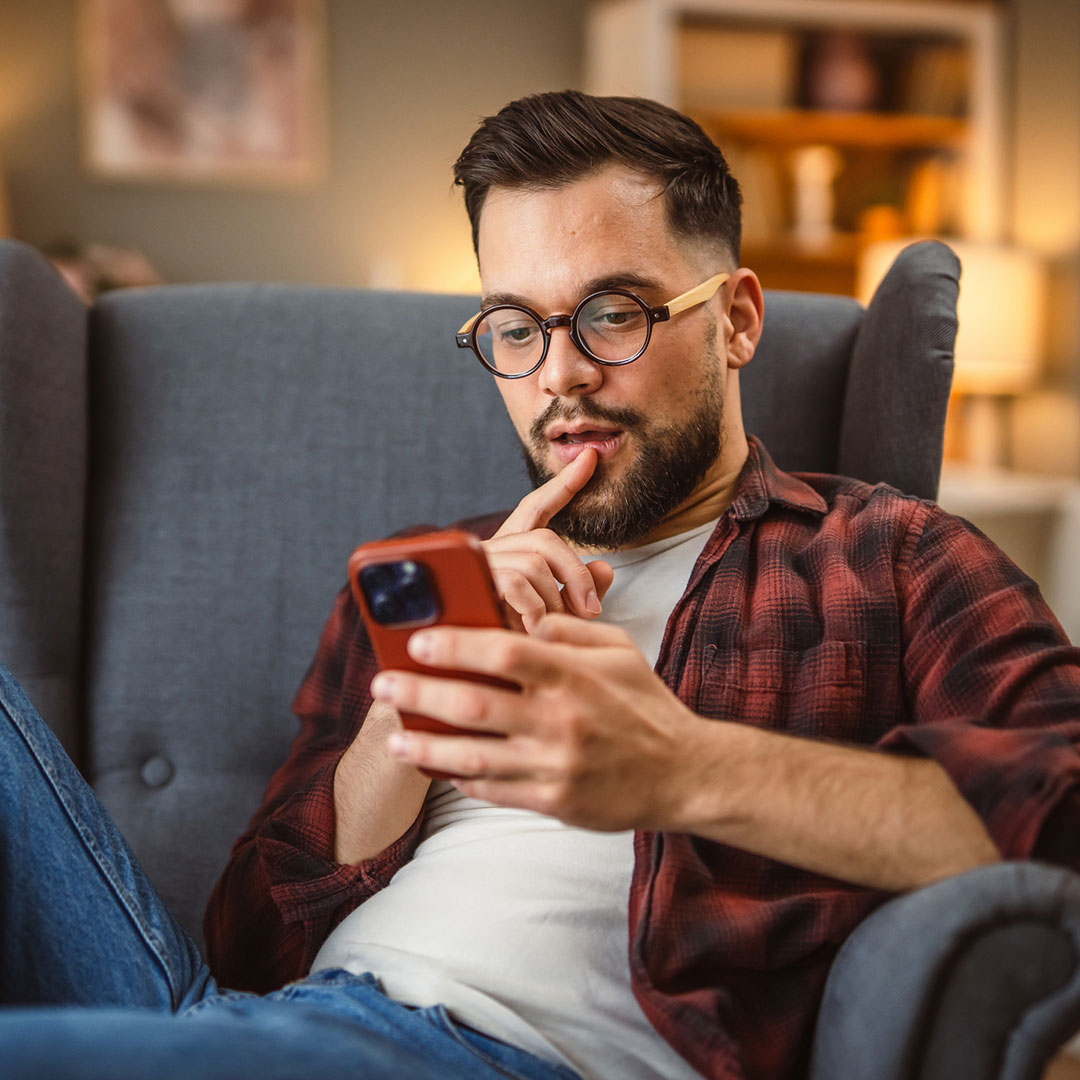 Photo of a man in an armchair in his home, reading on his mobile phone.