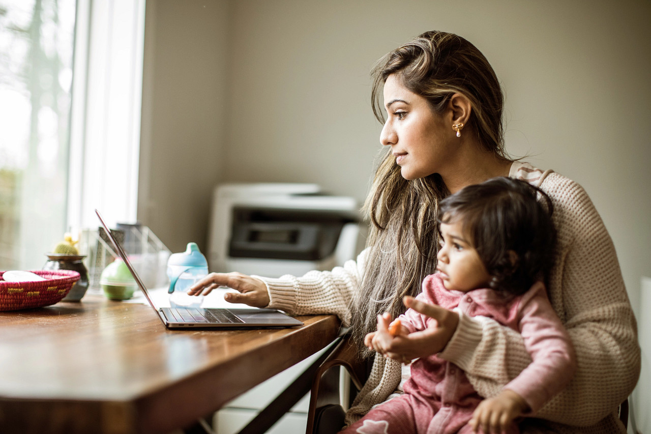 mother holding her young daughter while also working on a laptop at home