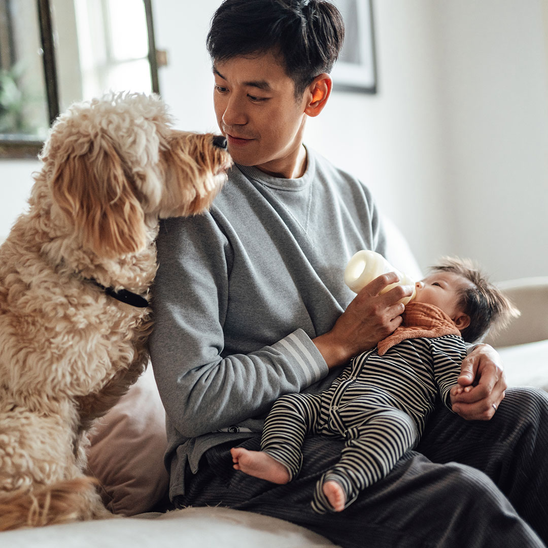 New father shows affection for the family dog while feeding his baby a bottle