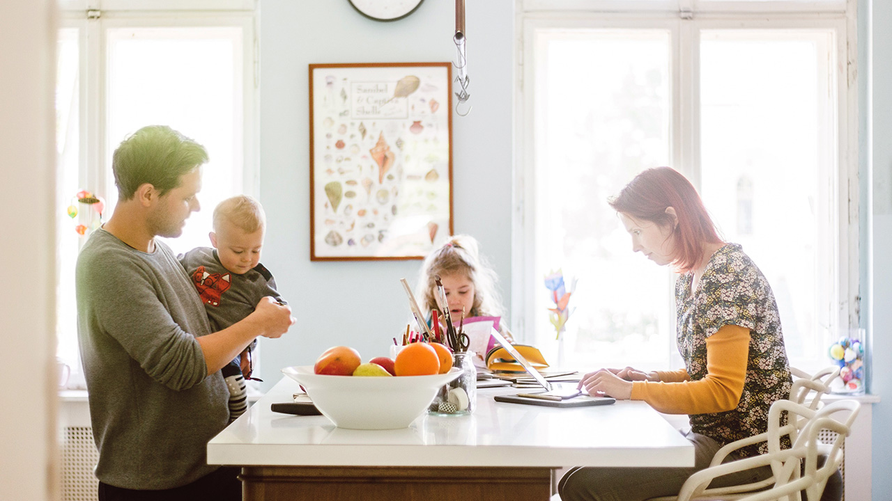 family of four gathered around the kitchen table while Mom seems to be working on her laptop